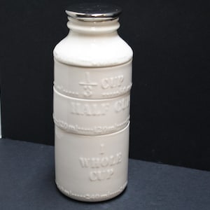 May include: A white ceramic measuring cup with a silver lid. The cup is cylindrical with embossed markings for 1/3 cup, half cup, and whole cup. The measurements are also listed in milliliters. A kitchenware item.