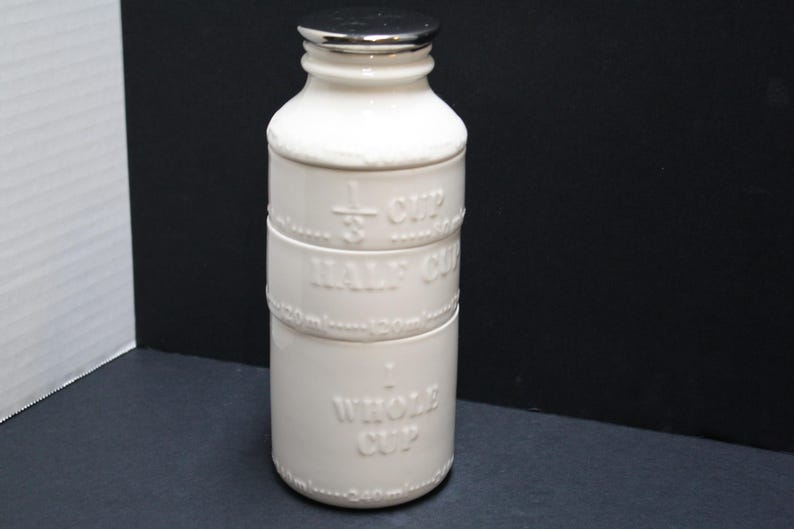 May include: A white ceramic measuring cup with a silver lid. The cup is cylindrical with embossed markings for 1/3 cup, half cup, and whole cup. The measurements are also listed in milliliters. A kitchenware item.