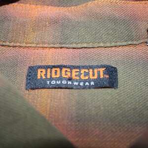 May include: A black label with white text that reads "Ridgecut Toughwear" on a green and orange plaid shirt.