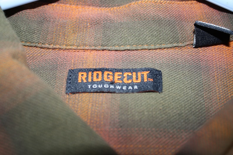 May include: A black label with white text that reads "Ridgecut Toughwear" on a green and orange plaid shirt.