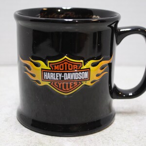 May include: Black ceramic mug with a curved handle. The mug features the Harley-Davidson logo in orange and yellow flames. The mug is a classic style, suitable for hot beverages.