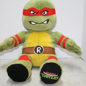 May include: Green Teenage Mutant Ninja Turtles plush toy with a red bandana and a yellow shell. The toy has a brown belt with a letter R on it. The toy is sitting on a white surface.