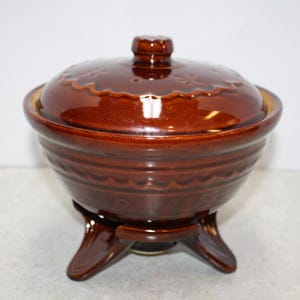 May include: A brown ceramic casserole dish with a lid. The dish has a round shape with a decorative pattern around the base and lid. It sits on three curved feet.