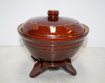 May include: A brown ceramic casserole dish with a lid. The dish has a round shape with a decorative pattern around the base and lid. It sits on three curved feet.