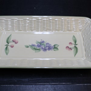 May include: A rectangular ceramic serving tray with a woven pattern and a green rim. The tray features a floral design with purple flowers and green leaves, and two pink cherries on each side.