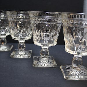 May include: Four clear, vintage-style stemmed glasses. Each glass features a square base, a decorative stem, and a detailed, textured bowl. The glasses are arranged in a row, showcasing their intricate design and clarity.