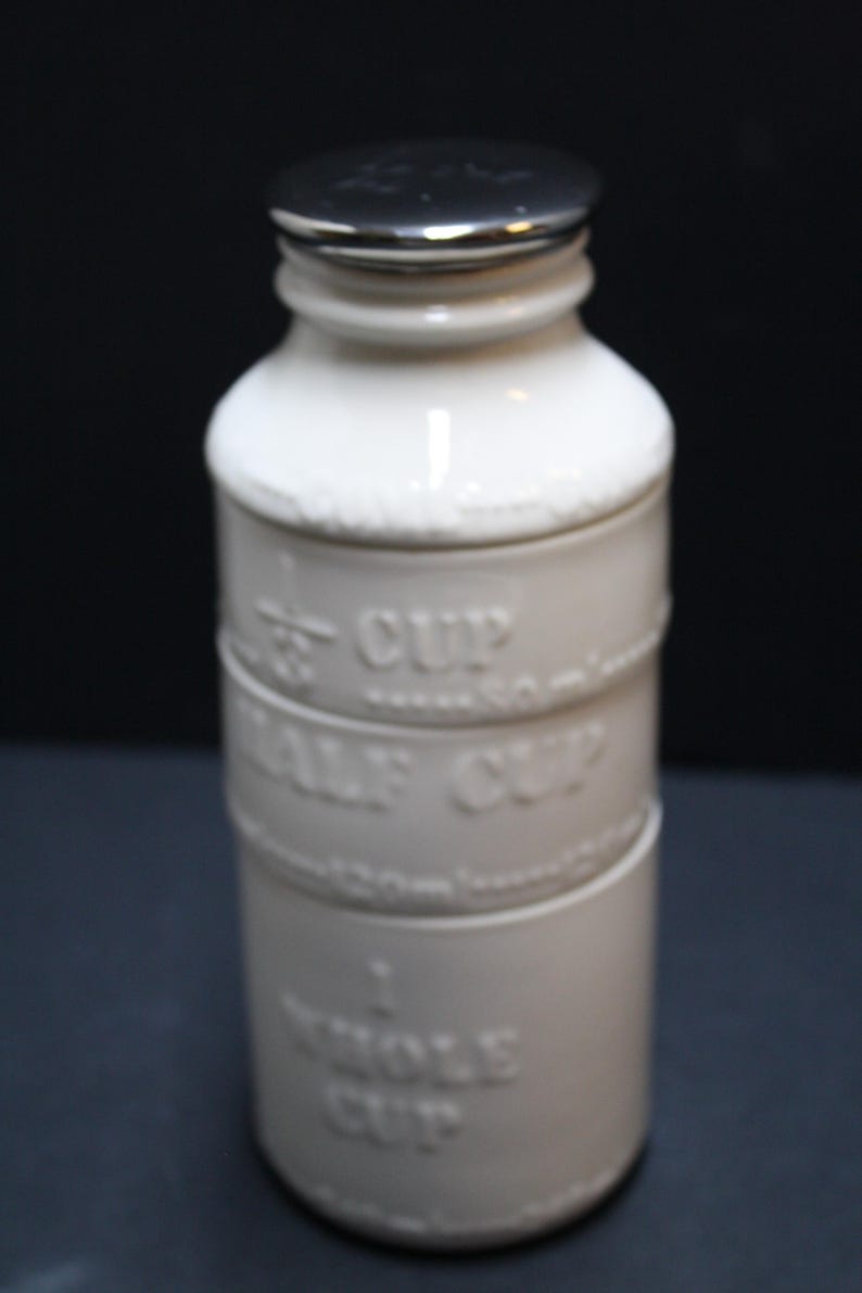 May include: A white ceramic measuring cup set with a silver lid. The set includes 1/3 cup, 1/2 cup, and 1 whole cup measurements. The embossed text is in a classic font, ideal for kitchen use.