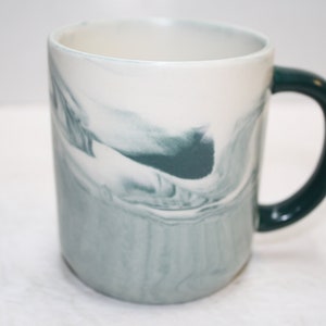 May include: A white ceramic mug with a teal marbled design and a dark teal handle.