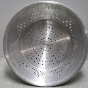 May include: A round, silver-colored metal colander featuring a brushed finish. The interior surface displays a series of small, circular drainage holes arranged in a central circular pattern, surrounded by six distinct triangular groupings of holes.