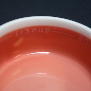 May include: Close-up of a white and coral measuring cup. The interior is coral, with the words "1/3 cup" and "80 ml" printed in white. The rim is white. The background is black.