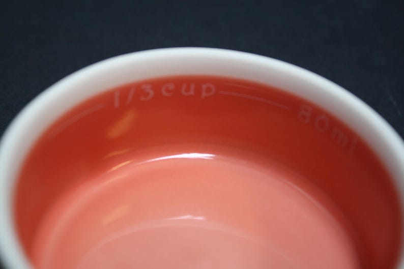 May include: Close-up of a white and coral measuring cup. The interior is coral, with the words "1/3 cup" and "80 ml" printed in white. The rim is white. The background is black.