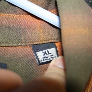 May include: A close-up of a brown, orange, and green plaid shirt with a black tag that reads "XL MADE IN BANGLADESH".