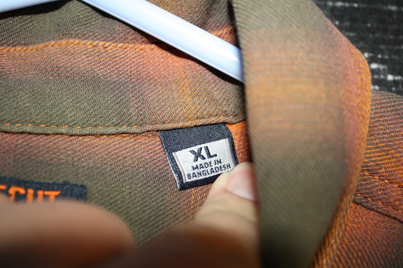 May include: A close-up of a brown, orange, and green plaid shirt with a black tag that reads "XL MADE IN BANGLADESH".