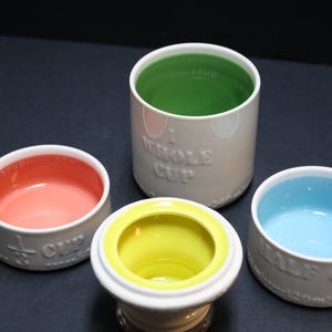 May include: A set of four ceramic measuring cups in various sizes. The cups are white with colorful interiors: pink, yellow, green, and blue. Each cup is labeled with its measurement, such as "1/2 CUP" or "WHOLE CUP".
