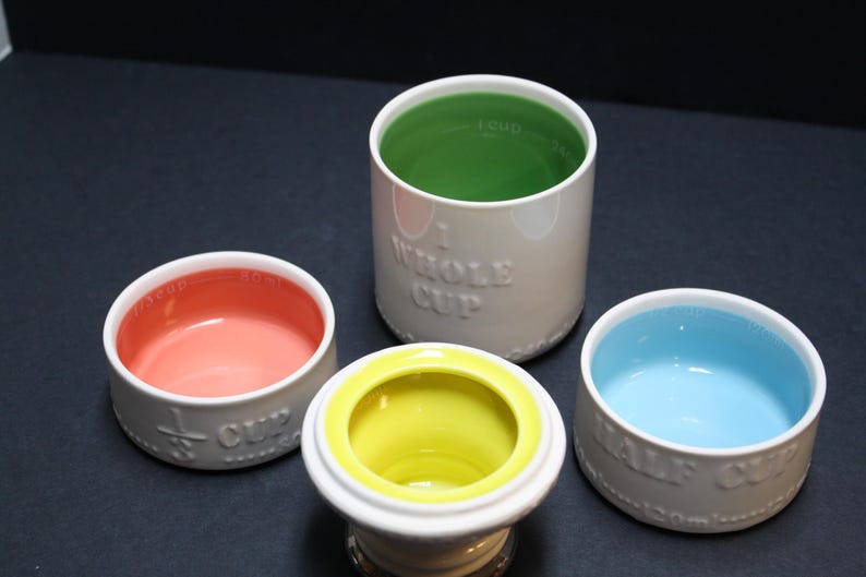 May include: A set of four ceramic measuring cups in various sizes. The cups are white with colorful interiors: pink, yellow, green, and blue. Each cup is labeled with its measurement, such as "1/2 CUP" or "WHOLE CUP".