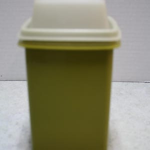 May include: A vintage olive green rectangular container with a cream-colored lid. The container has a square shape and a slightly textured surface. The lid fits snugly on top, creating a tight seal. This storage container is ideal for kitchen or pantry organization.