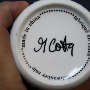May include: White ceramic mug base with black text that reads "made in china", "fabriqu&eacute; en chine", and "dishwasher safe". The mug features a signature in black ink, "R Costa".