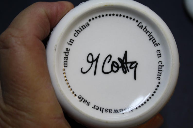 May include: White ceramic mug base with black text that reads "made in china", "fabriqu&eacute; en chine", and "dishwasher safe". The mug features a signature in black ink, "R Costa".