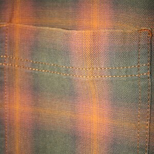 May include: Close-up of a brown and orange plaid fabric with a sewn pocket. The fabric has a subtle diagonal weave.