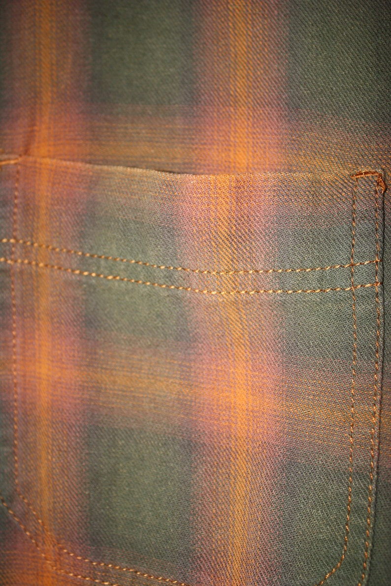 May include: Close-up of a brown and orange plaid fabric with a sewn pocket. The fabric has a subtle diagonal weave.