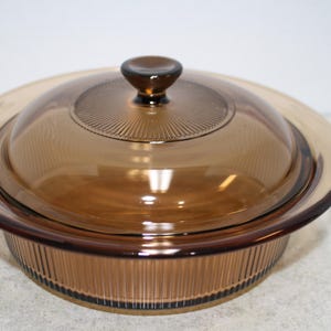 May include: A round, amber-colored glass casserole dish with a matching lid. The dish has a ribbed exterior and a smooth, clear lid with a central knob. The lid has a decorative, ridged pattern. The dish is designed for cooking and serving.