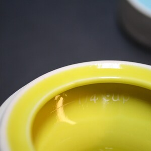 May include: Close-up of a white and yellow ceramic measuring cup. The interior is bright yellow, with the words "1/4 cup" etched into the side. The rim is white, and the background is a dark gray.