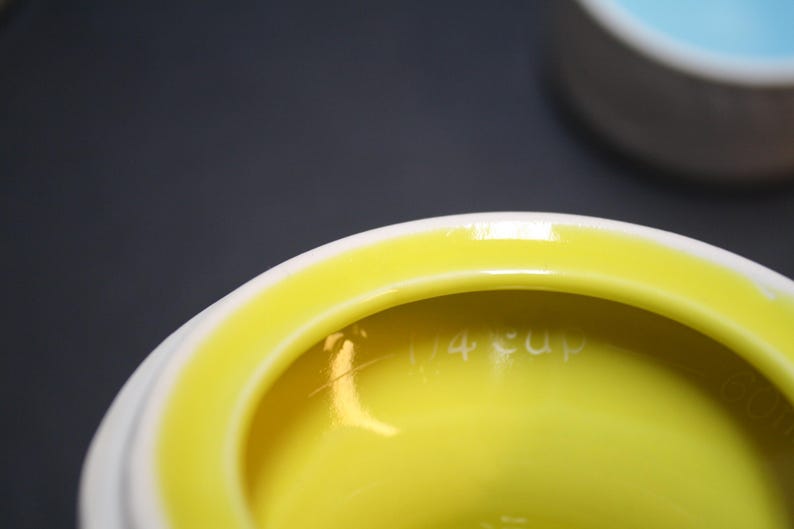 May include: Close-up of a white and yellow ceramic measuring cup. The interior is bright yellow, with the words "1/4 cup" etched into the side. The rim is white, and the background is a dark gray.