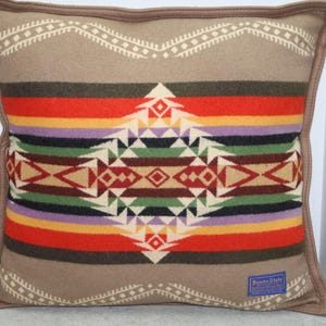 May include: A square throw pillow featuring a tan base with a central geometric pattern in red, orange, purple, green, and black. The pillow has a brown piped edge and a small blue rectangular tag in the bottom right corner with the text Beaver State.