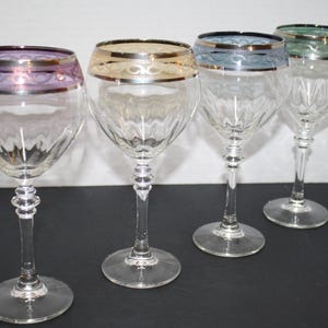 May include: Four clear wine glasses with decorative rims in purple, gold, blue, and green. Each glass has a clear, patterned bowl and a detailed stem. The glasses are arranged in a row, showcasing their elegant design.