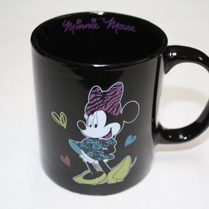 Puede incluir: Taza de cer&aacute;mica negra con un gr&aacute;fico de Minnie Mouse con un atuendo de estampado de cebra y un lazo rosa. La taza tiene el texto "Minnie Mouse" en morado en el borde.