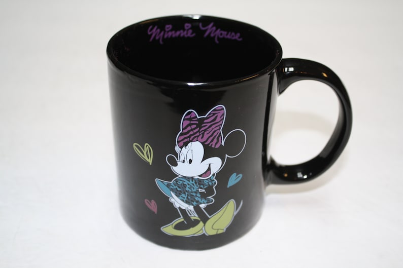 Puede incluir: Taza de cer&aacute;mica negra con un gr&aacute;fico de Minnie Mouse con un atuendo de estampado de cebra y un lazo rosa. La taza tiene el texto "Minnie Mouse" en morado en el borde.