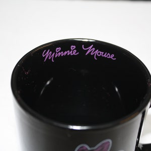 Puede incluir: Taza de caf&eacute; de cer&aacute;mica negra con una firma de Minnie Mouse en color morado y dos corazones en el borde interior.