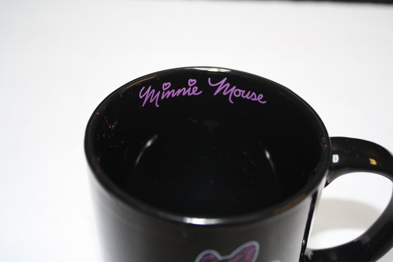 Puede incluir: Taza de caf&eacute; de cer&aacute;mica negra con una firma de Minnie Mouse en color morado y dos corazones en el borde interior.