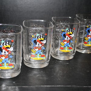 May include: Set of four clear glass Mickey Mouse cups with a colorful design featuring Mickey Mouse dressed as a conductor. The cups have a 1990s Disney theme and feature the text "1990-1991 Disney's Hollywood Studios 10th Anniversary".