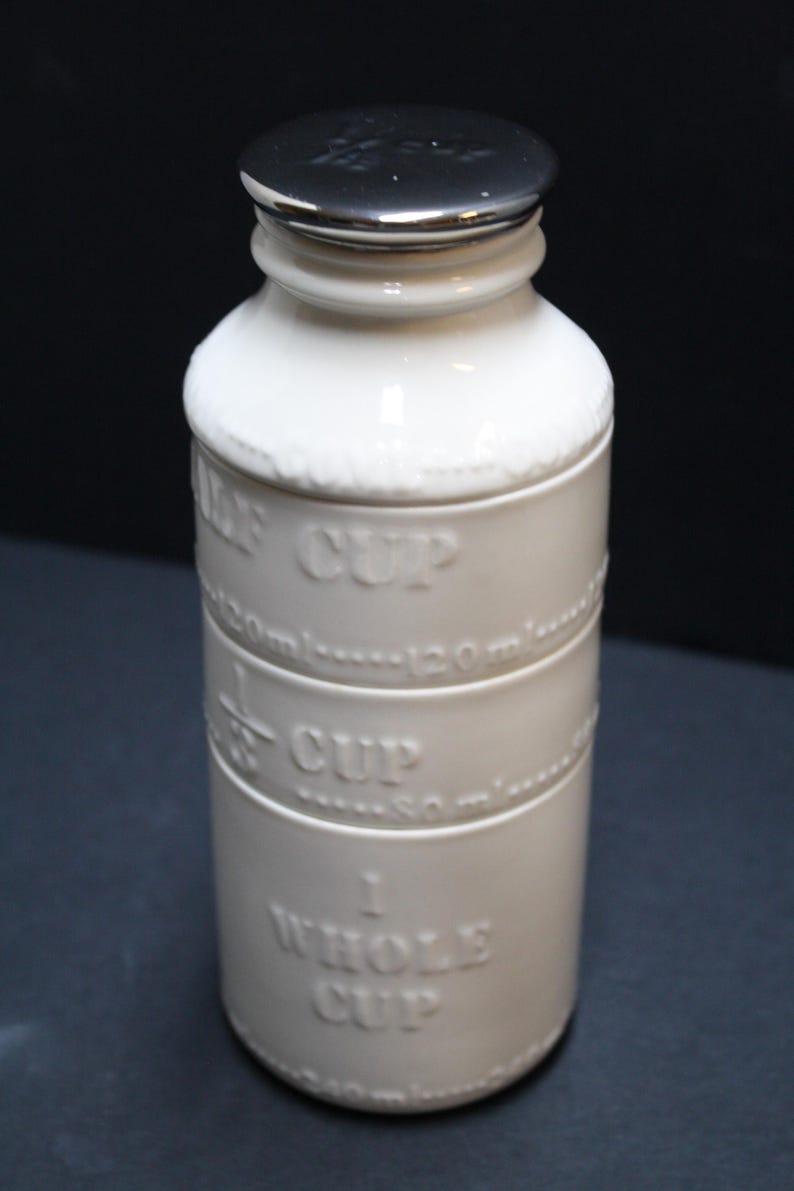 May include: A white ceramic measuring cup with a silver lid. The cup has embossed markings for 1/2 cup, 1/3 cup, and 1 whole cup, with corresponding measurements in milliliters. The design is simple and functional.