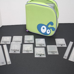 May include: A green carrying case with a blue ant design and the text "Cuttlebug" on the front. The case is open and contains a set of clear plastic embossing folders with the text "Cuttlebug" on each.