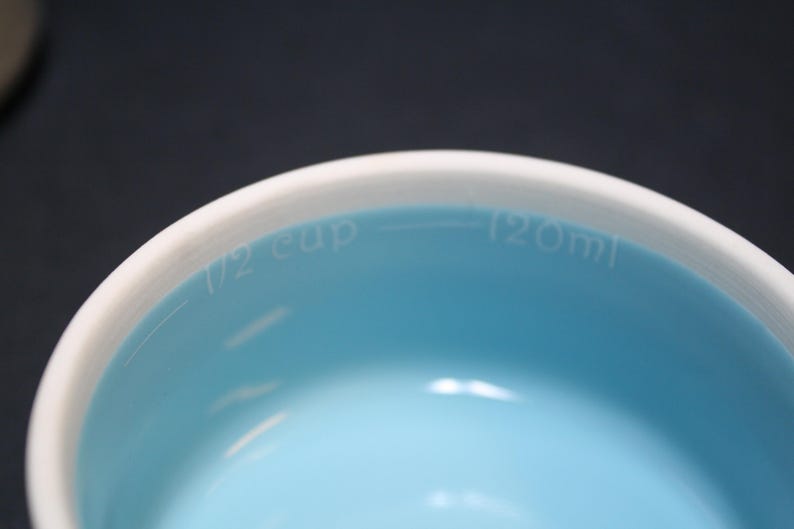 May include: Close-up of a measuring cup with a white rim and a light blue interior. The text "1/2 cup" and "120ml" are printed on the inside of the cup, indicating the measurement capacity. The cup is designed for measuring liquid or dry ingredients.