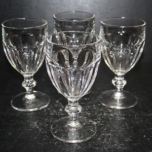 May include: Four clear crystal wine glasses with a decorative, cut-glass design. Each glass has a round base, a detailed stem, and a flared bowl. The glasses are arranged on a dark surface, showcasing their intricate patterns and clarity.