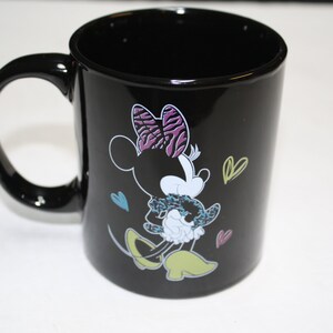 Puede incluir: Taza de caf&eacute; de cer&aacute;mica negra con una ilustraci&oacute;n colorida de Minnie Mouse con un lazo morado y blanco y sosteniendo una Minnie Mouse m&aacute;s peque&ntilde;a. La taza tiene un asa blanca y est&aacute; decorada con peque&ntilde;os corazones.