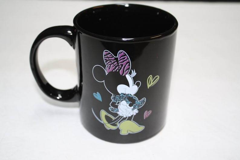 Puede incluir: Taza de caf&eacute; de cer&aacute;mica negra con una ilustraci&oacute;n colorida de Minnie Mouse con un lazo morado y blanco y sosteniendo una Minnie Mouse m&aacute;s peque&ntilde;a. La taza tiene un asa blanca y est&aacute; decorada con peque&ntilde;os corazones.