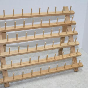 May include: A wooden thread rack with multiple rows of pegs. The rack is made of light-colored wood and has a tiered design, perfect for organizing spools of thread. The pegs are evenly spaced.
