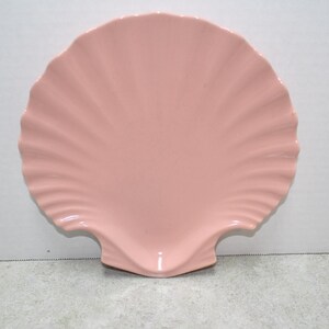 May include: A light pink, ceramic seashell-shaped dish. The dish has a scalloped edge and a smooth, glossy surface. The dish is designed to resemble a seashell, with a fan-like pattern radiating from the center.