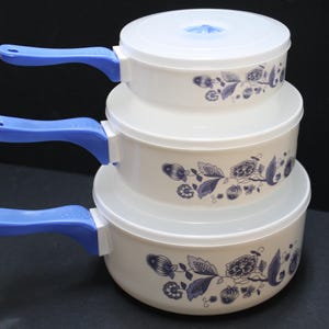 May include: A set of three nesting pots with lids. The pots are off-white with a floral design in blue. Each pot has a blue handle. The lids are white. The pots are stacked on top of each other.