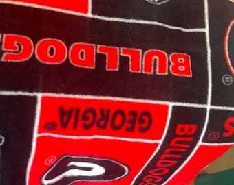 May include: A red, black, and white fleece blanket featuring the word "BULLDOG" in red block letters. The blanket also displays the word "GEORGIA" and the Bulldogs logo. The blanket has a black border.