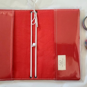 May include: A red fabric notebook cover with clear vinyl pockets on the interior flaps, featuring white elastic cords for securing inserts. A small rectangular label with text is sewn onto the right side. Two rolls of patterned tape are placed nearby.
