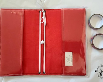 Puede incluir: Una funda de tela roja para cuaderno con bolsillos de vinilo transparente en las solapas interiores, con cordones elásticos blancos para asegurar los insertos. Una pequeña etiqueta rectangular con texto está cosida en el lado derecho.