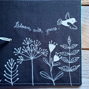 Peut inclure: Un carnet en tissu noir avec une broderie blanche de fleurs, de feuilles et d'un oiseau. Le texte "Bloom with grace" est brod&eacute; sur la couverture.