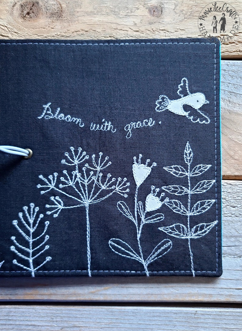 Peut inclure: Un carnet en tissu noir avec une broderie blanche de fleurs, de feuilles et d'un oiseau. Le texte "Bloom with grace" est brod&eacute; sur la couverture.