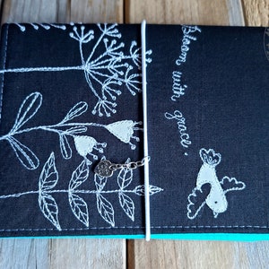 Peut inclure: Journal en tissu noir avec broderie blanche de fleurs et d'un oiseau. Le journal a une fermeture en ruban blanc et un charme argent&eacute;. Le texte "Bloom with grace." est brod&eacute; sur la couverture.