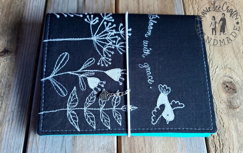 Peut inclure: Journal en tissu noir avec broderie blanche de fleurs et d'un oiseau. Le journal a une fermeture en ruban blanc et un charme argent&eacute;. Le texte "Bloom with grace." est brod&eacute; sur la couverture.